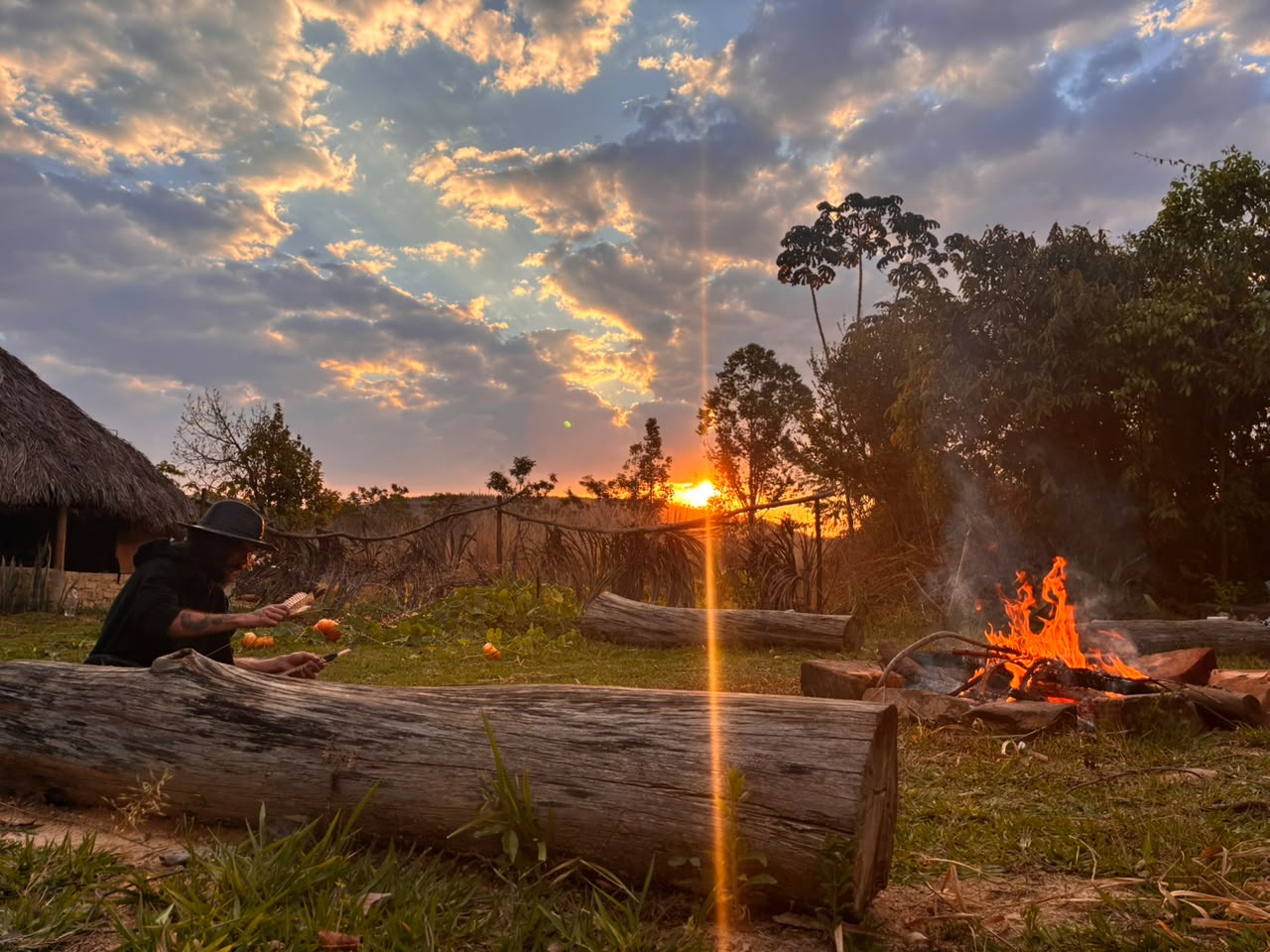Fogueira acesa e pôr do sol sobre a oca de palha do retiro